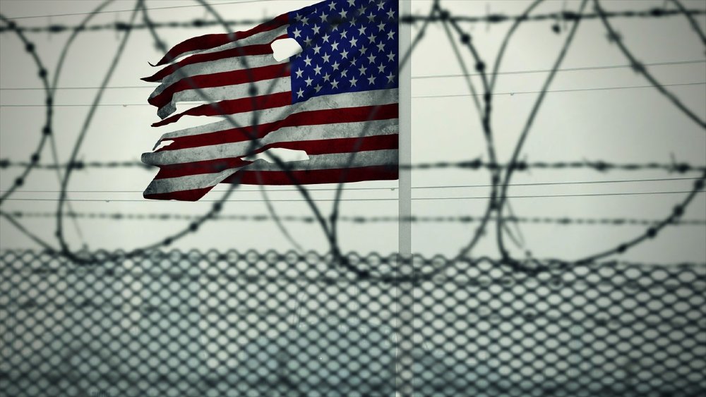 Photo of a barbed wire fence with a tattered American flag flying in the background
