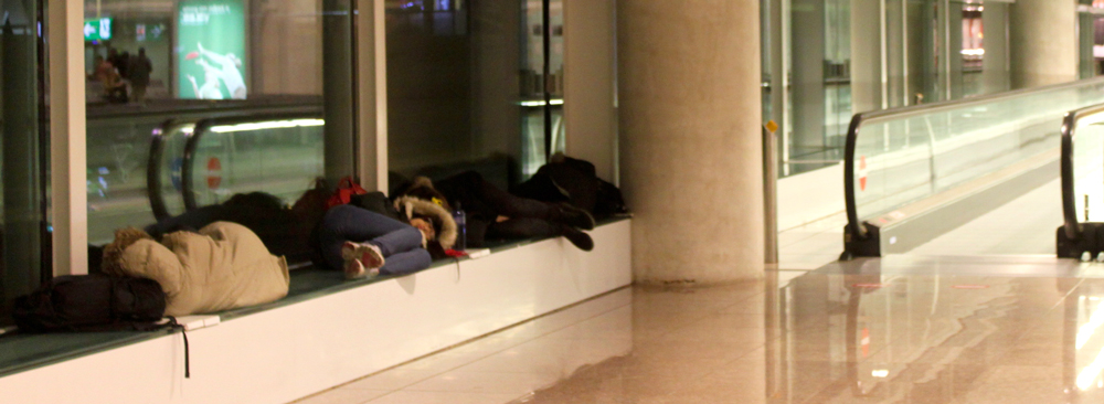 Photo showing people curled up on a ledge in an airport next to a motorized walkway.
