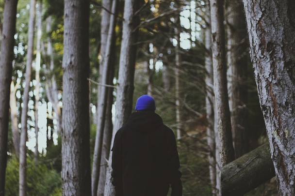 A man wearing a black coat and blue cap walks in the woods.