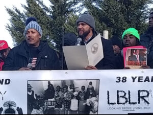 Photo of three men, one holding a microphone and read off a paper.