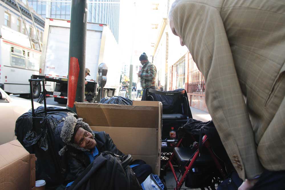Phillip Alston leans over a woman, bundled in warm clothes and laying down amid cardboard boxes, surrounded by tattered luggage and a walker.