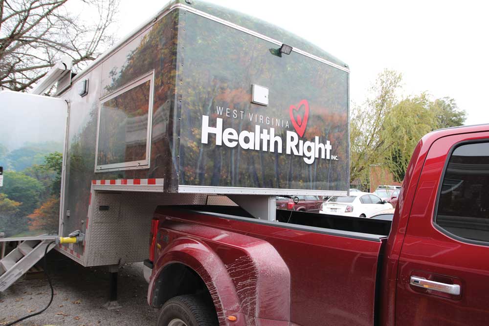 A trailer, labeled West Virginia Health Right, with an open door and steps leading inside, hooked into a red pickup truck with mud around the wheels. There are trees in the background.