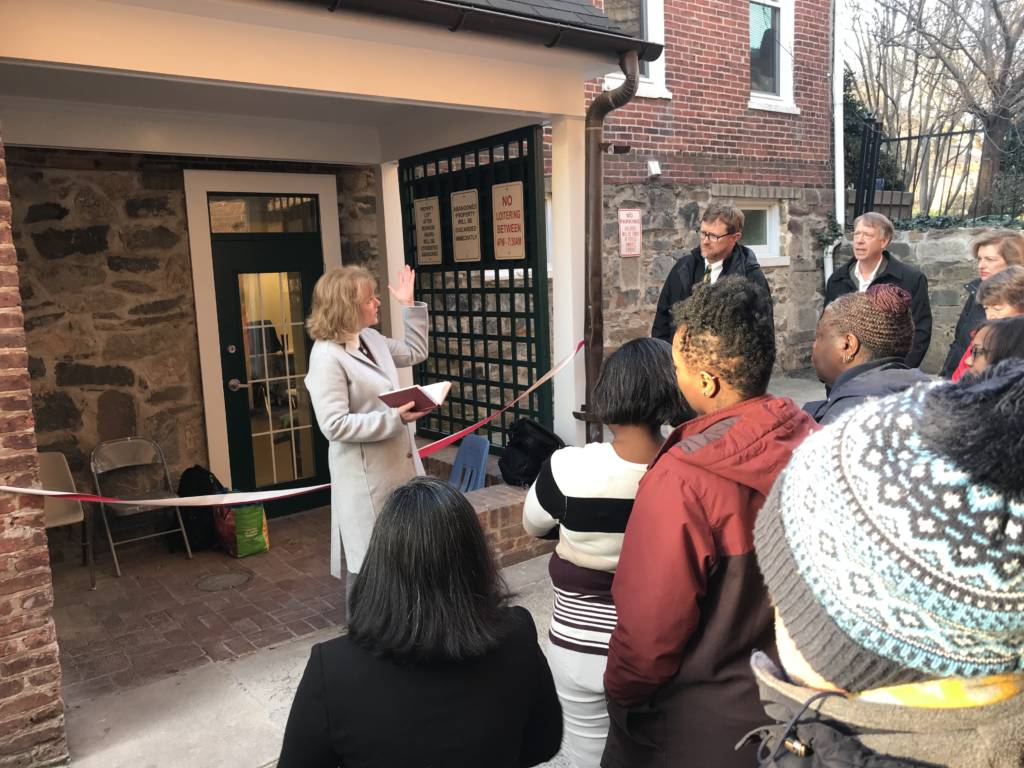 Outdoors in an alley, a minister reads from a holy book and holds up a hand to bless a crowd.