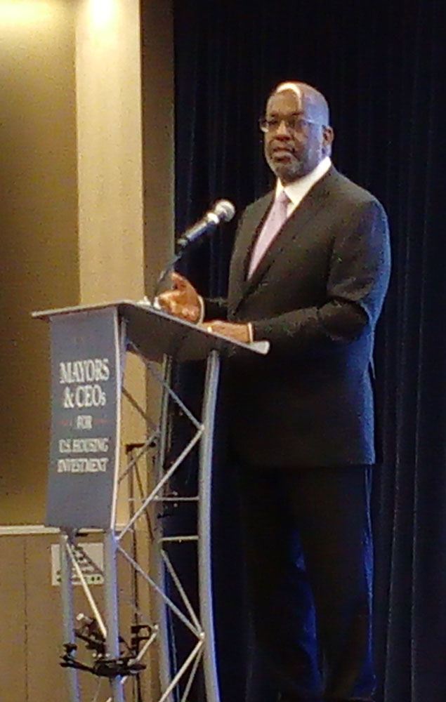 Photo of Bernard Tyson, wearing a suit, standing at a podium with the label "Mayors and CEOs for U.S. Housing Investment