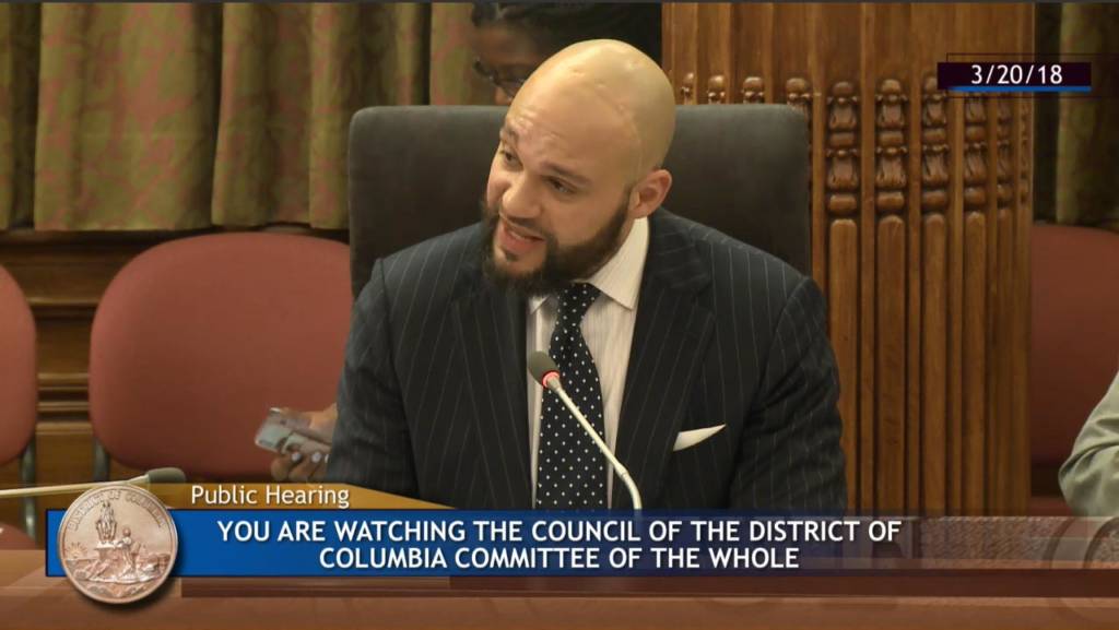 Screenshot of a man speaking into a microphone. Text on the screen says "You are watching the council of the district of columbia committee of the whole" and displays the date "03/20/2018"