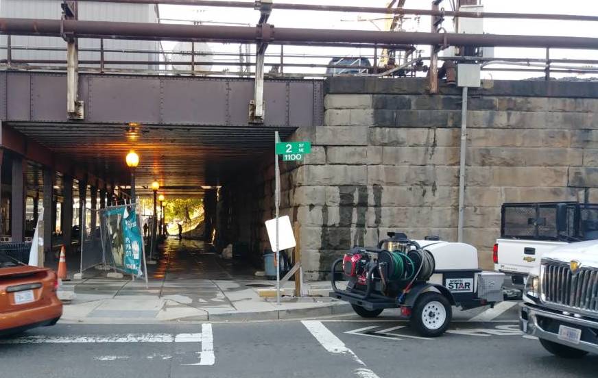 A photo showing an underpass with a "2nd Street NE" sign in front of it at the intersection. A parked truck with a water trailer sits next to it and the sidewalk in the underpass is wet. A fence is closing off the sidewalk, but the side of it is open where the pressure-washing had been taking place.