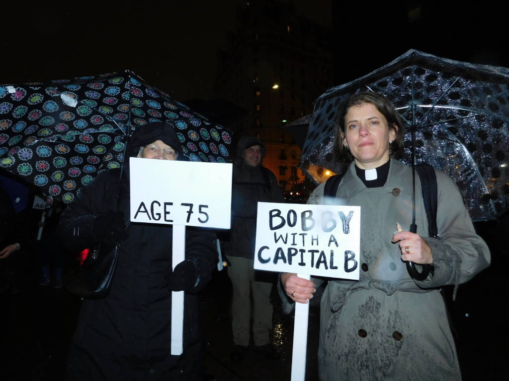 Photo of two women, each using an umbrella, and carrying a sign to represent one of the people who died while homeless.