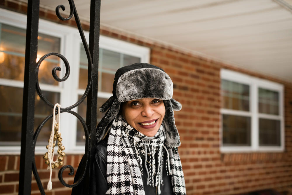 Color photo of a woman in a winter hat, scarf, and coat leaning against a decorative metal post in front of a brick house