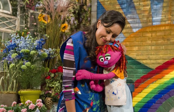 Pink, Sesame Street Muppet hugs woman in front of colorful brick wall.