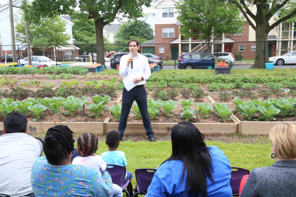 Photo of a man standing in front of planting beds, speaking to a seated crowd.