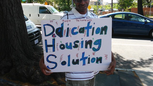 A man holding a sign that reads "Dedicated Housing Solution" at a protest.