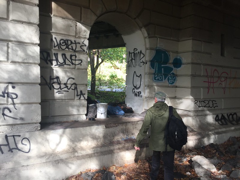 Photo of a man with a backpack standing under a bridge, approaching someone who appears to be sleeping.