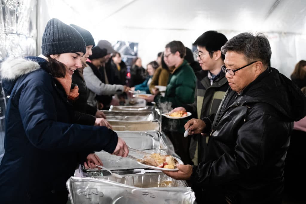 Photo of food trays with volunteer servers on one side and people receiving full plates on the other.