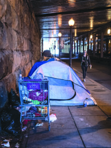 Caucasian woman holding metal water bottle and wearing headphones walks by tent and shopping cart of belongings owned by homeless resident of NoMa underpass.