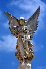 A plaster statue of an angel with large wings holding a cross. The background is a blue sky with scattered clouds with