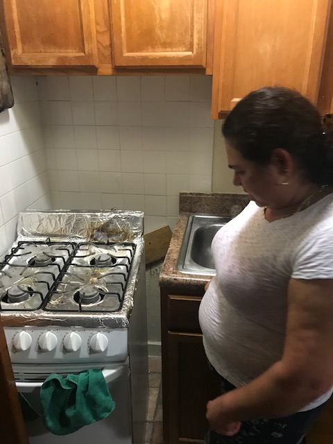 A hispanic woman stands next to her stove pulled away from the wall to display a piece of wood nailed over a hole.