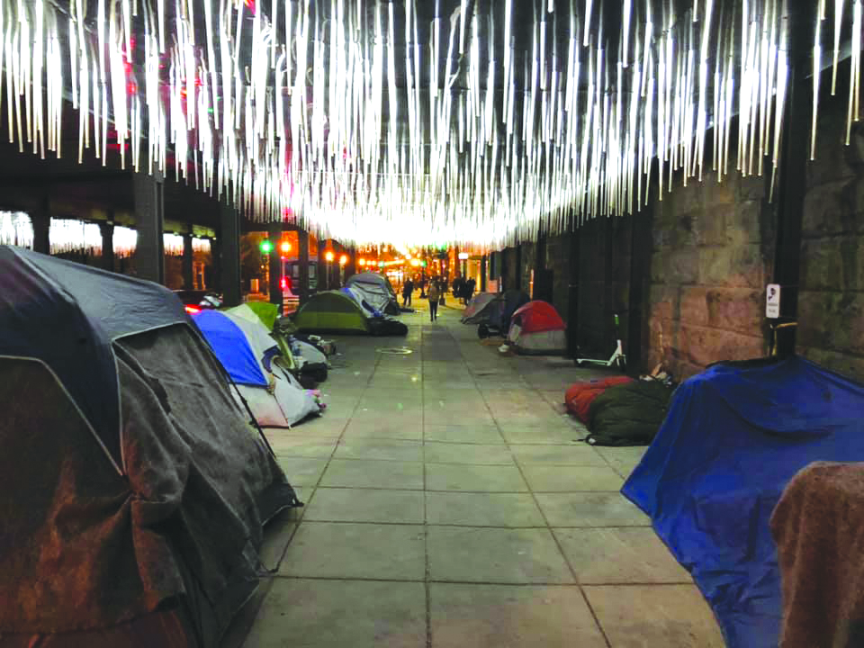 Photo showing tubes of light in a tunnel over a wide sidwalk where tents have been pitched.