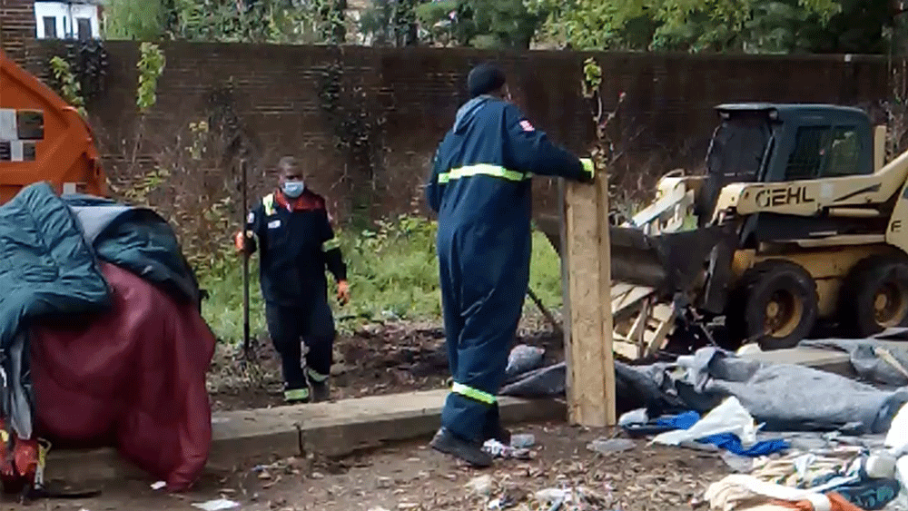 Two workers in jumpsuits and wearing masks clear debris between a garbage truck and a skid loader.