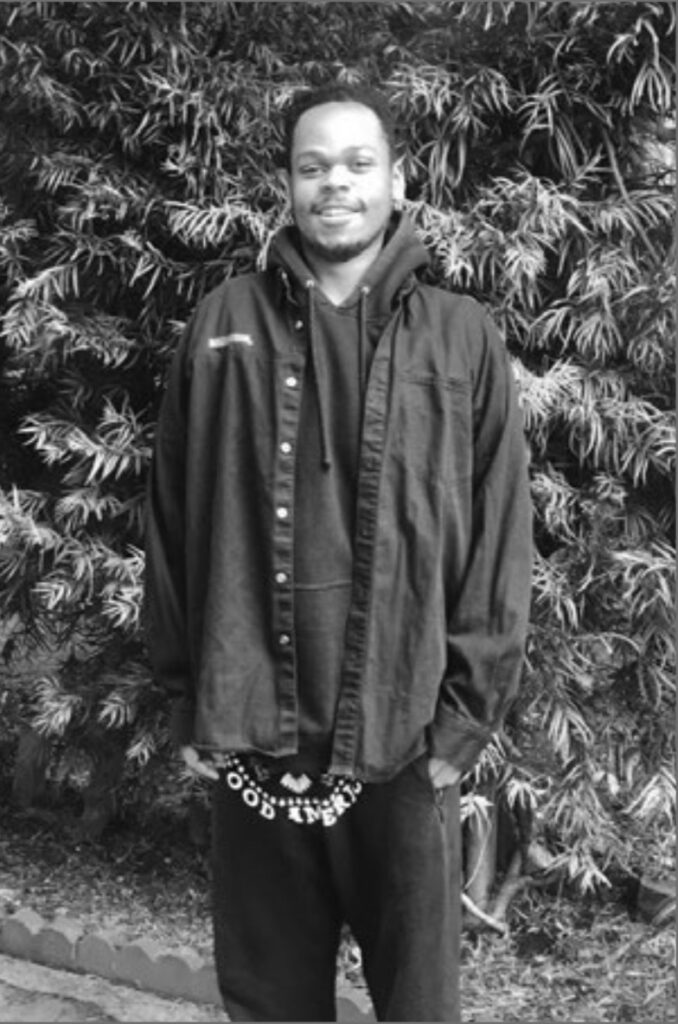 A black young man stands in front of a bush. He wears a black sweatshirt and sweatpants and a loose button up.