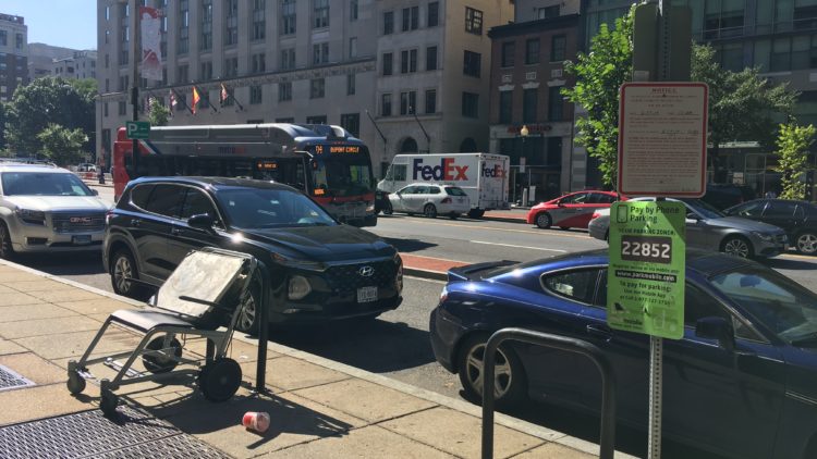 A D.C. city street. A red and white sign advertises an upcoming encampment clean-up. Cars are parked on the side of the road and a bus is passing by in the photo.