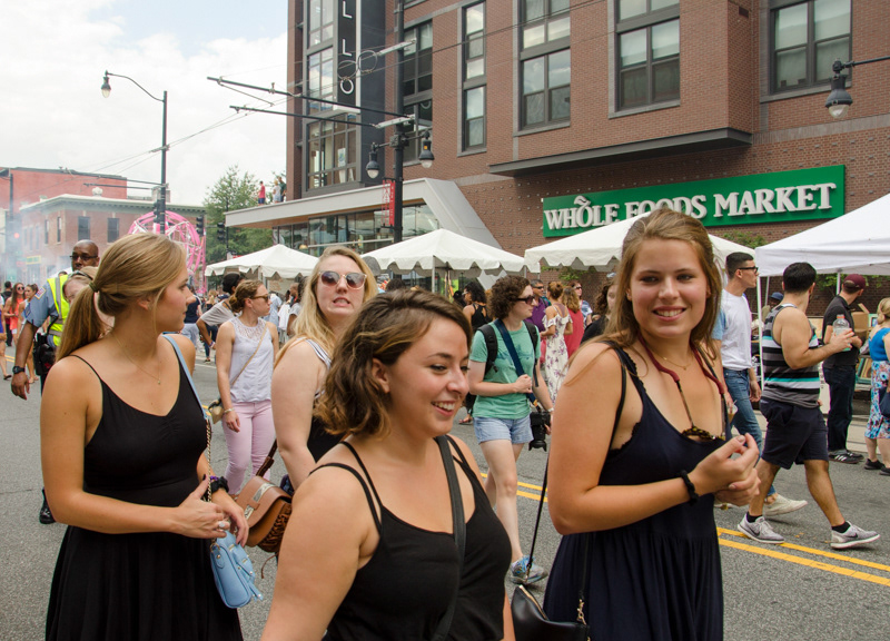 Four white girls in black dresses smile and walk through a street market. A green Whole Foods sign is behind them.