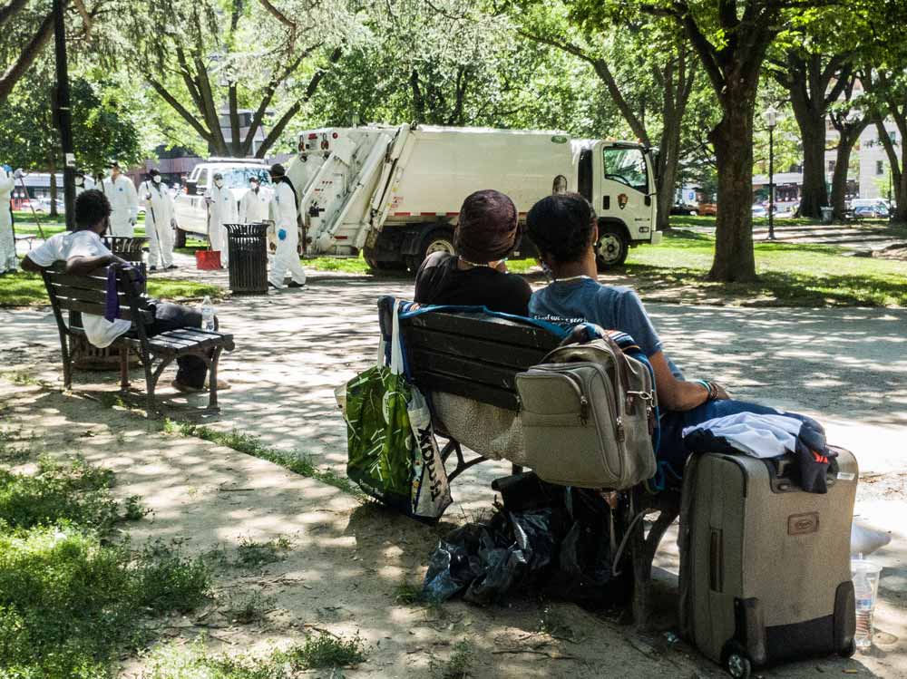 Photo of two people on a bench, one with many belonging, watching clean up workers wearing protective white suits, standing next to and a garbage truck.