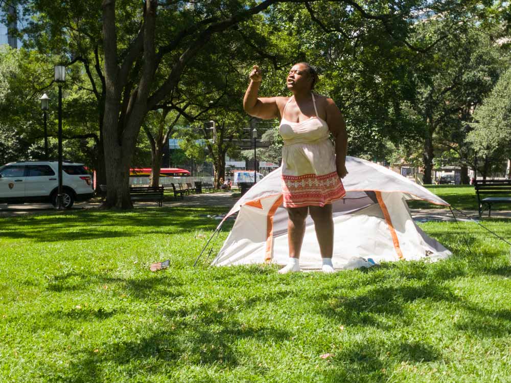 Photo f a woman standing up in front of her tent, pointing and speaking emphatically.