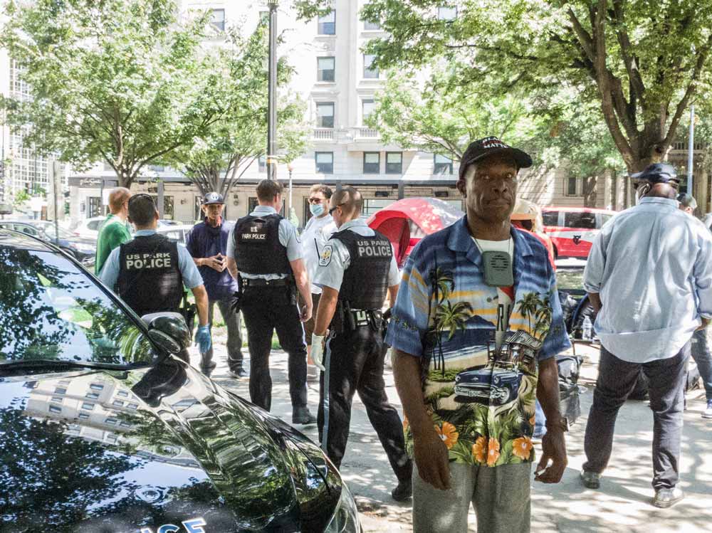 Photo of a man in a "Black Lives Matter" hat looking directly at the camera. U.S. Park Police officers are visible behind him.