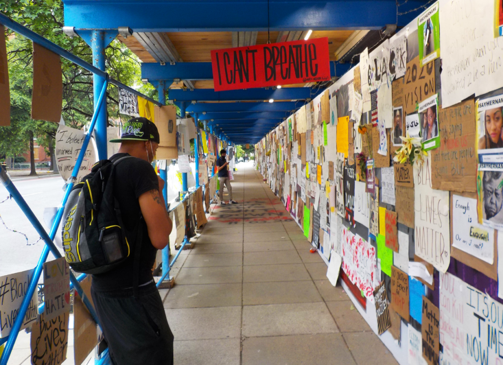 Photo of a man looking at many posters tacked onto a temporary wall under scaffolding. One prominent orange sign reads "I can't breathe." 
