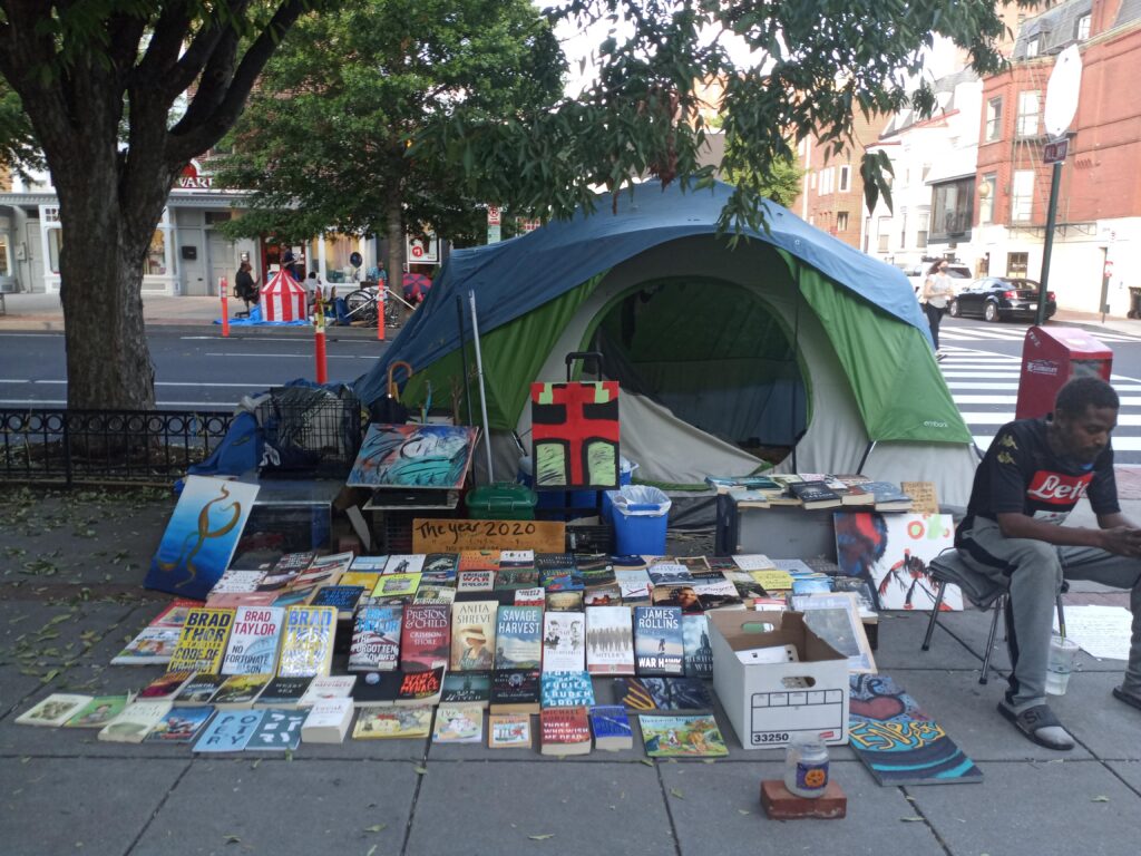Photo showing a man sitting on a chair in front of a green and gray tent. Books, artwork, and a tip jar are displayed on the sidewalk next to him in front of the tent.