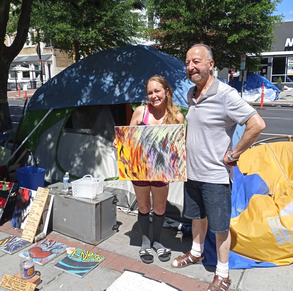 Photo showing a woman holding an abstract painting and standing next to an older man, both smiling at the camera.