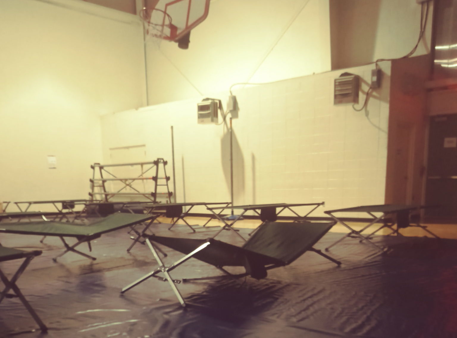 Photo of six empty cots in the gym, where plastic has been put down across the gym floor. A basketball goal is visible above.