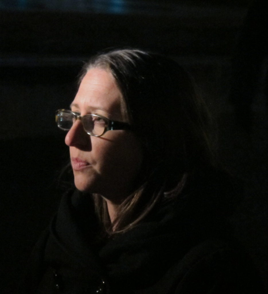 A photo taken at night shows only a woman's face, dramatically lit. Her dark hair and dark coat melt into her surroundings.