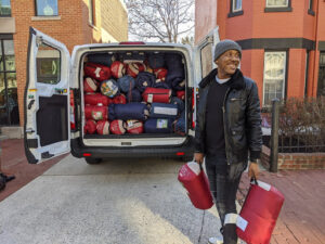 Image shows Kirk “Scooby” Ellis holding two red sleeping bags in front of a van with it's truck open, showing stacks of sleeping bags. 