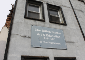 Photos displays The Mitch Snyder Art & Education Center for the Homeless, a white building with a blue sign and two windows
