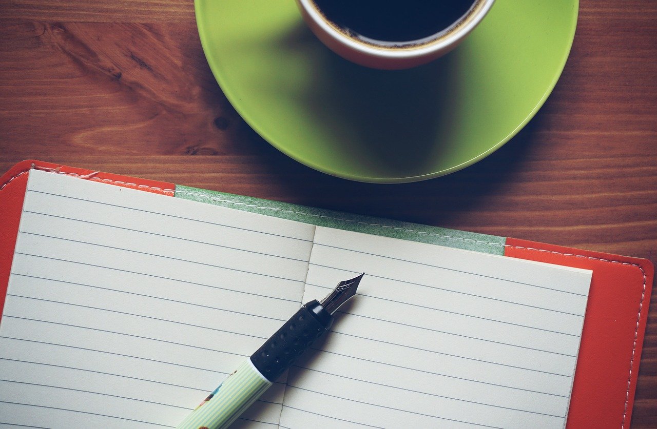 Photo of a cup of coffee, a blank notebook, and a fountain ink pen.
