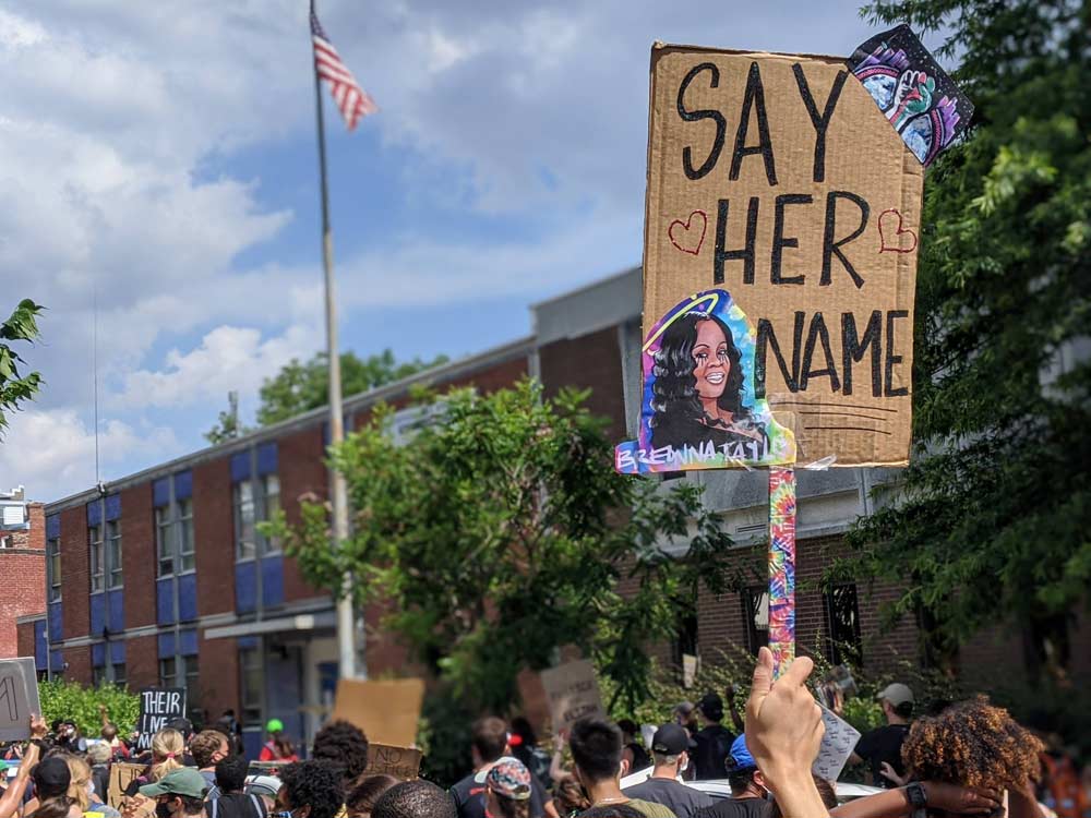 Photo of a sign that reads "Say Her Name" and depicts Breonna Taylor is held above a crowd outside of a police station.