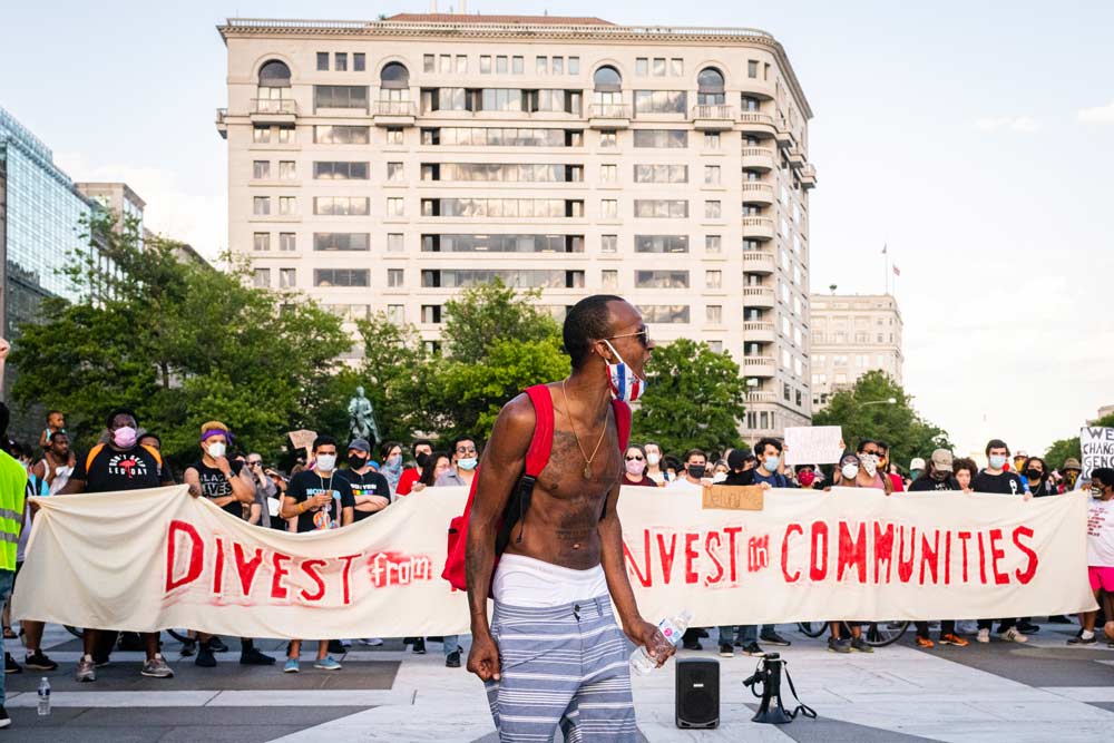 Photo of a man chanting during a racial justice demonstration. A crowd behind him carries a banner that says "divest from police, invest in communities"