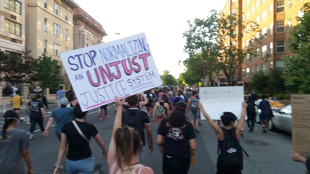Photo taken amid a racial justice march. A woman is holding up a sign that says "stop normalizing an unjust justice system."