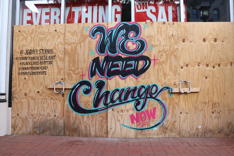 Mural painted on plywood that is sealing a closed business; the mural says "We Need Change Now"