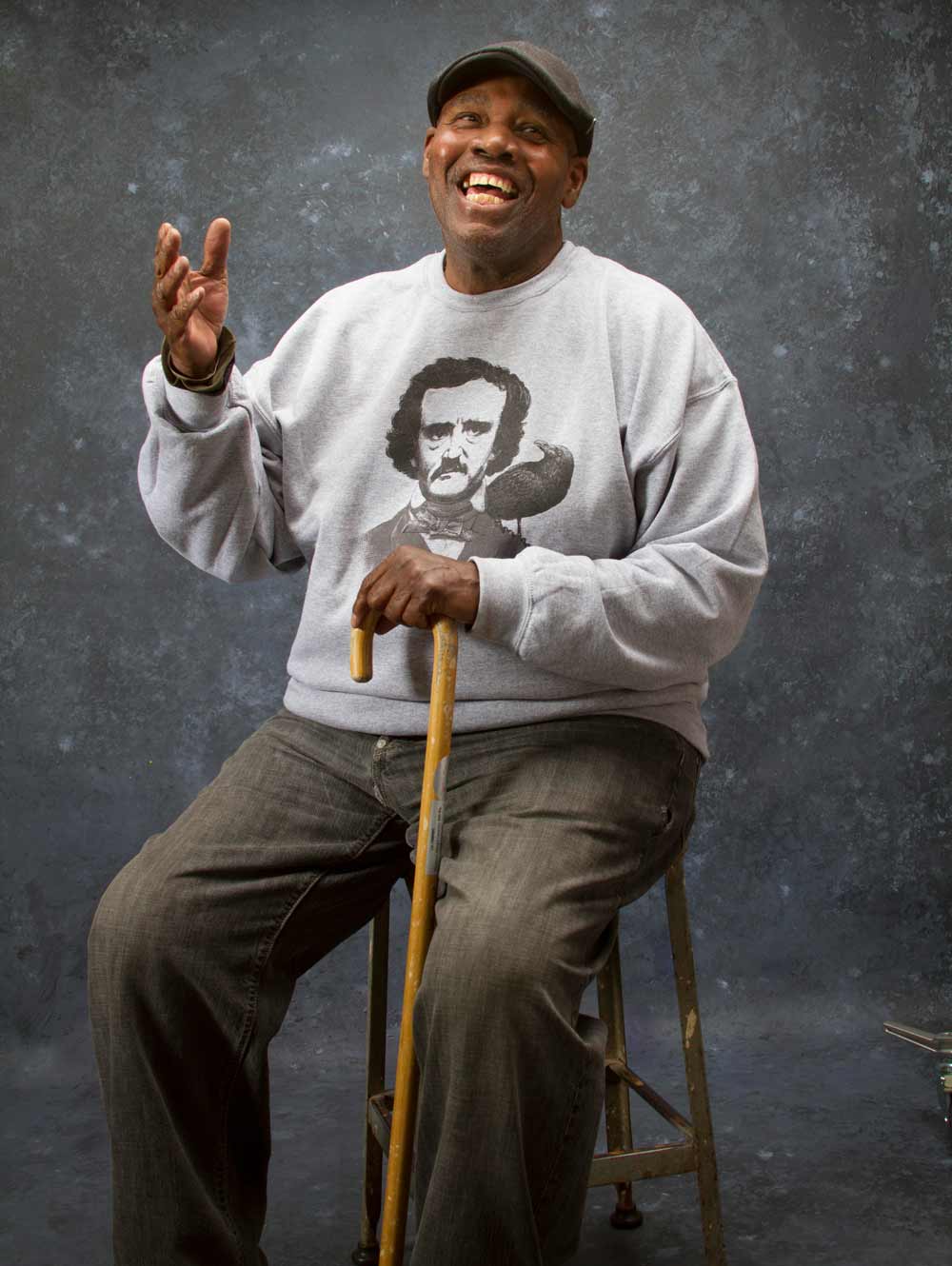 Photo of David seated on a stool with a dark backdrop behind him. He's laughing, wearing an Edgar Alan Poe sweatshirt and holding his cane.