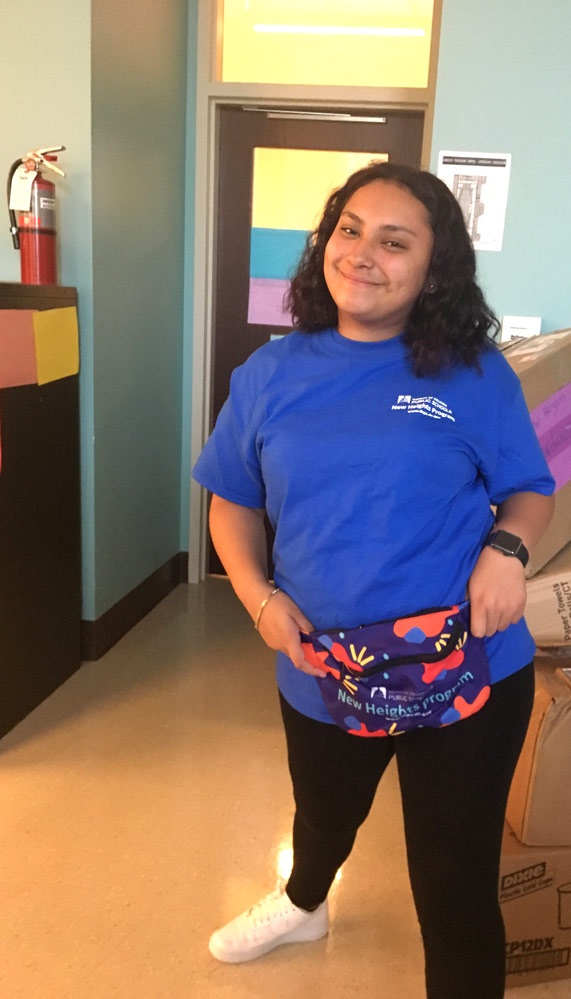 Photo of a smiling young woman wearing a shirt that says "DC Public Schools" and a multi-colored bag that says "New Heights" 