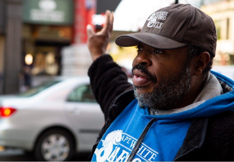 A man raises his hand as he offers to sell wares on the sidewalk.