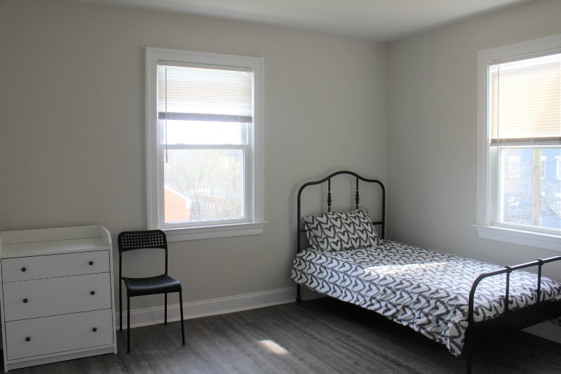 A full size bed is pictured, as well as bedroom furniture such as a dresser and extra chair. The room is well lit.