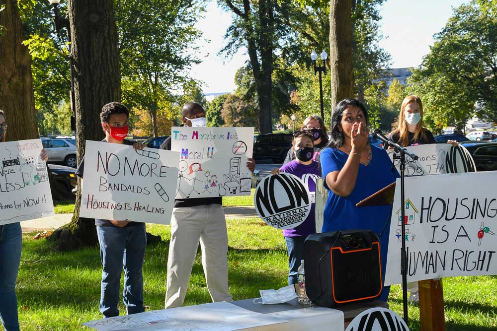 Photo of a woman speaking at a podium while others behind her hold signs reading "No more band-aids, housing is healing," "the moratorium isn't moral," etc.