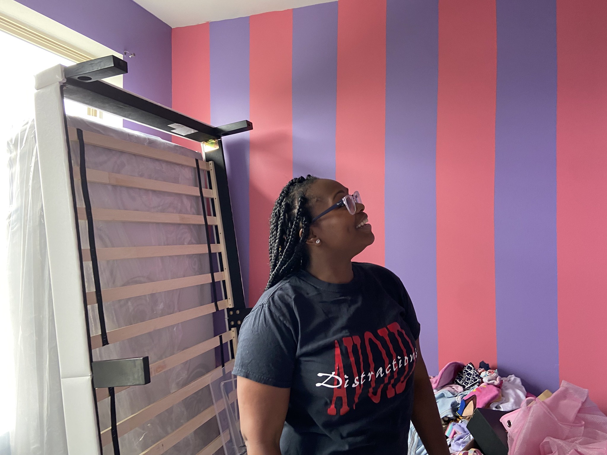 A woman looks up at the purple and pink striped walls of the room. A bed frame is pushed up against the wall behind her. 