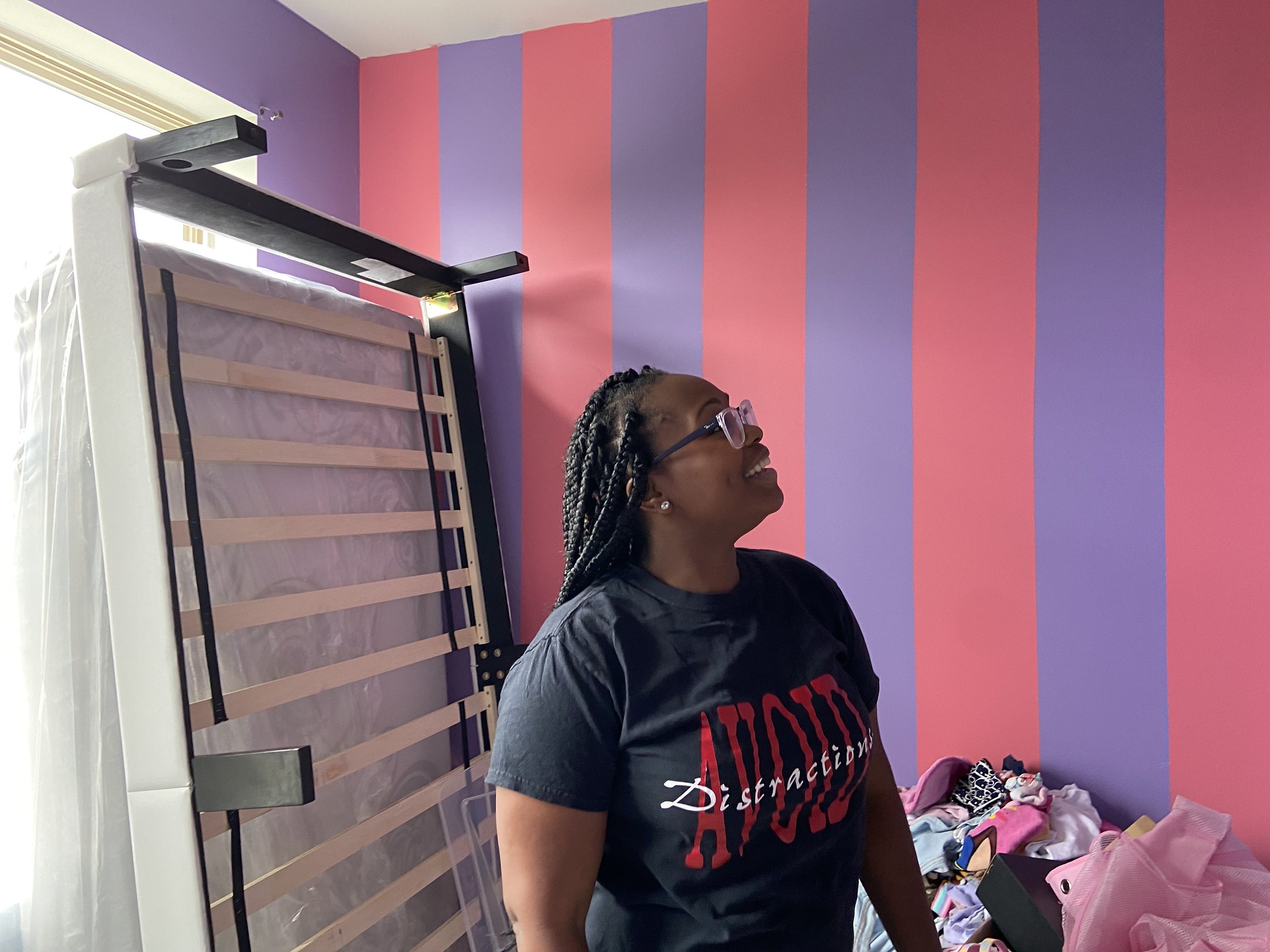 A woman looks up at the purple and pink striped walls of the room. A bed frame is pushed up against the wall behind her.