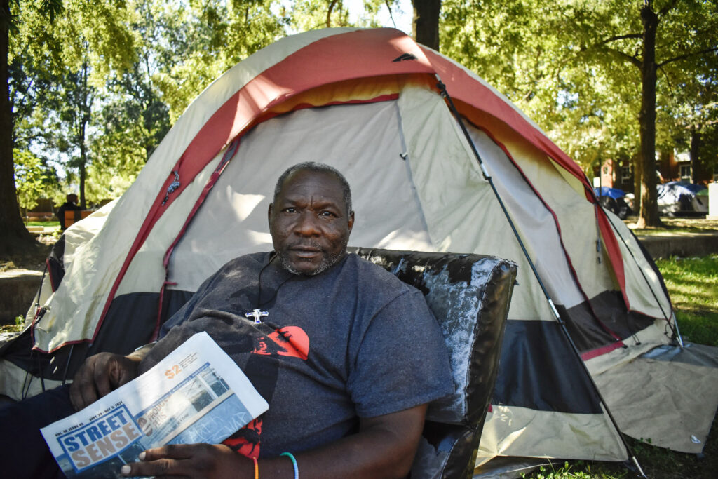 Aldo Richardson sits on his office chair in front of his tan and red tent at the New Jersey Ave. and O Street NW park, holding a Street Sense Media newspaper.