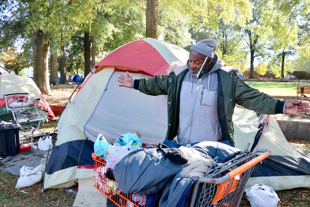 Photo of a man gesturing widely to a cart full of belongings. A tent can be seen behind him