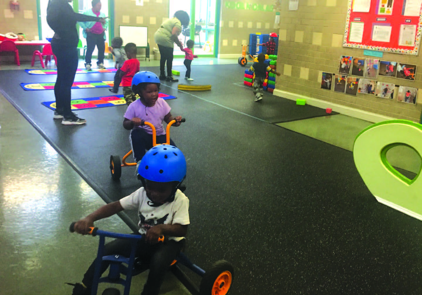 Photo of children pedaling small tricycles indoors on a playmat. Other children and adults are interacting in the background.
